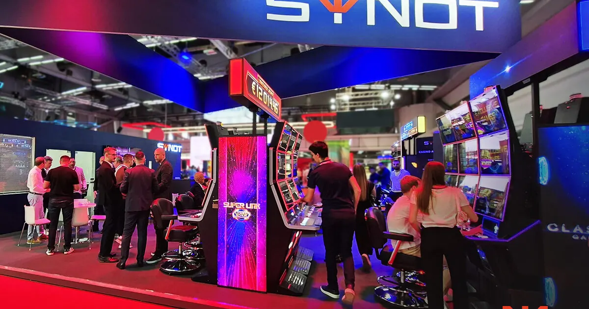 Um estande da SYNOT Group na feira Belgrade Future Gaming, exibindo gabinetes de jogos e telas com soluções omnicanal.