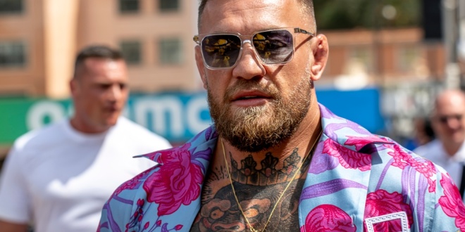 Conor McGregor sorrindo em um evento, com um logotipo discreto da 1xBet ao fundo.