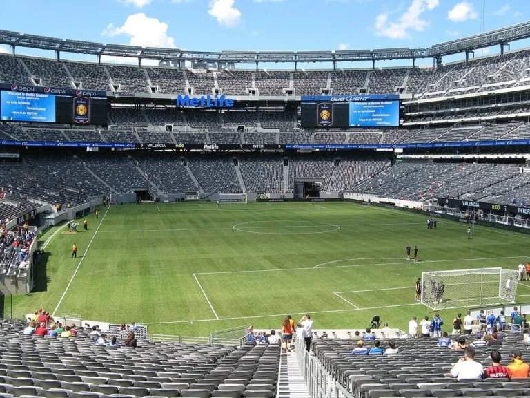 Uma visão aérea de um estádio de futebol lotado durante a final da Copa do Mundo, com telões mostrando replays e gráficos de jogo, e a marca FOX sutilmente visível.